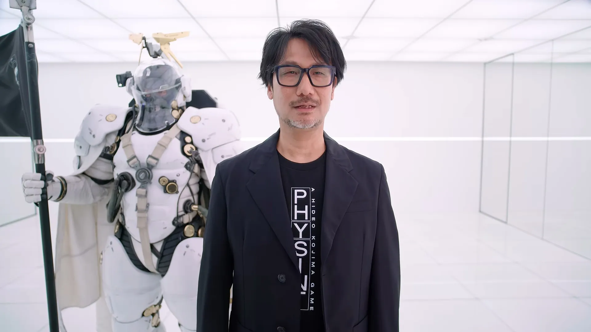 Hideo Kojima in a black blazer and t-shirt stands in a white room next to a person wearing a futuristic white armored suit holding a black flag.