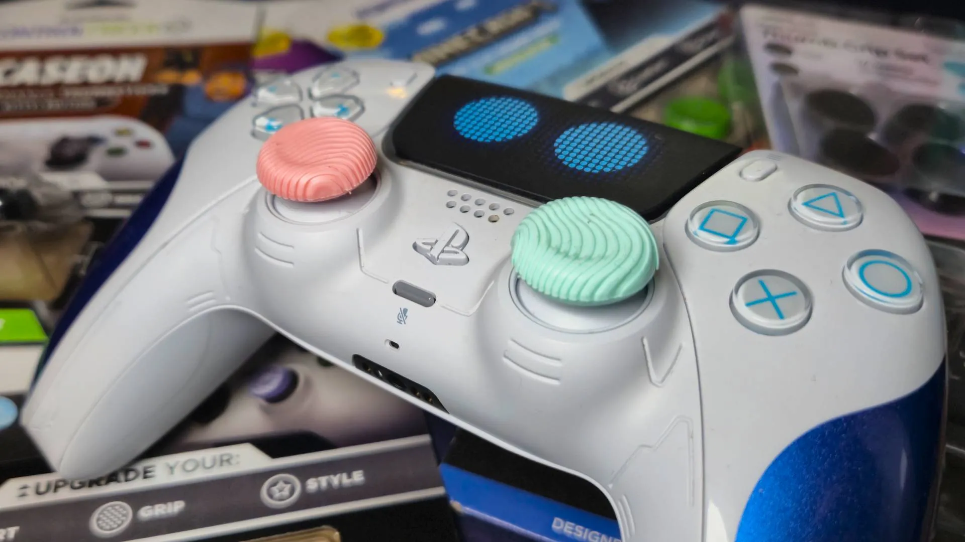 Photo of the Astro Bot PS5 DualSense controller with a pair of pink and green thumb grips. The controller is sitting on a pile of thumb grips packaging.